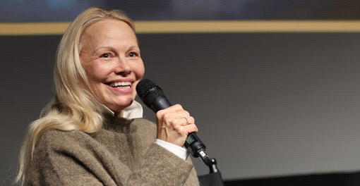 Pamela Anderson in a brown sweater, smiling while speaking into a microphone at an event