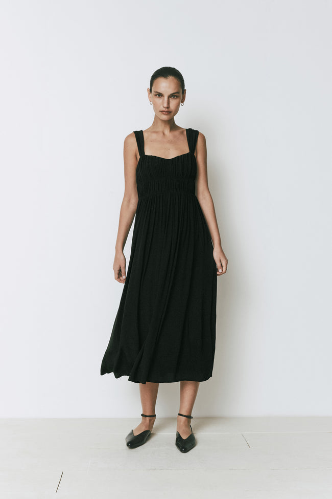 Black A-line midi Coucou Dress with gathered bodice, soft sweetheart neckline, and wide shoulder straps on a woman.