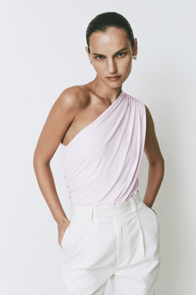 Perle Bodysuit in pink with ruched detail, one shoulder design, styled with white trousers.