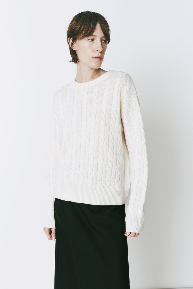 Ivory cable knit sweater with a crewneck, styled on a woman. Features wool-cashmere blend with a relaxed fit.