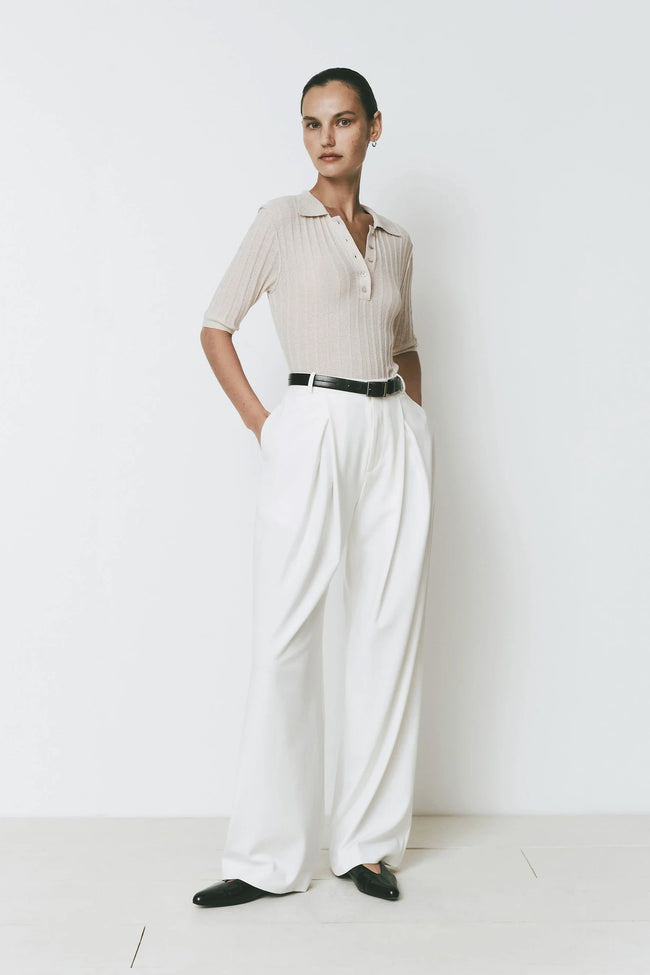 Woman wearing a cream polo shirt and white Drapé Pant with black belt and shoes, mid rise front pleated detail