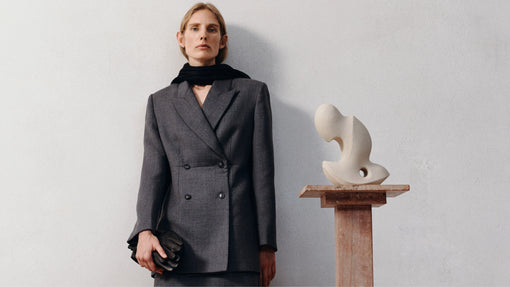 A woman in a grey double-breasted blazer and black scarf stands next to an abstract sculpture against a white wall