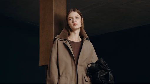 A fashion model poses in a beige coat with a corduroy collar, holding a large black leather bag against a dark background