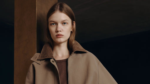 A young woman with blue eyes models a beige jacket with a brown corduroy collar, looking directly at the camera