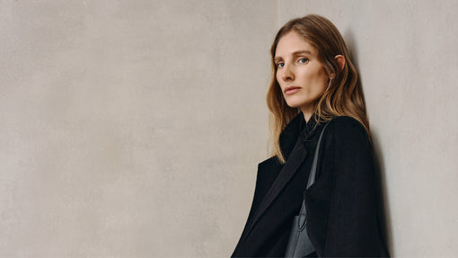A woman with long wavy hair wearing a black coat leans against a wall, looking seriously at the camera