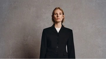 A woman stands against a grey background, wearing a black blazer, wide-leg blue jeans, and holding a black tote bag
