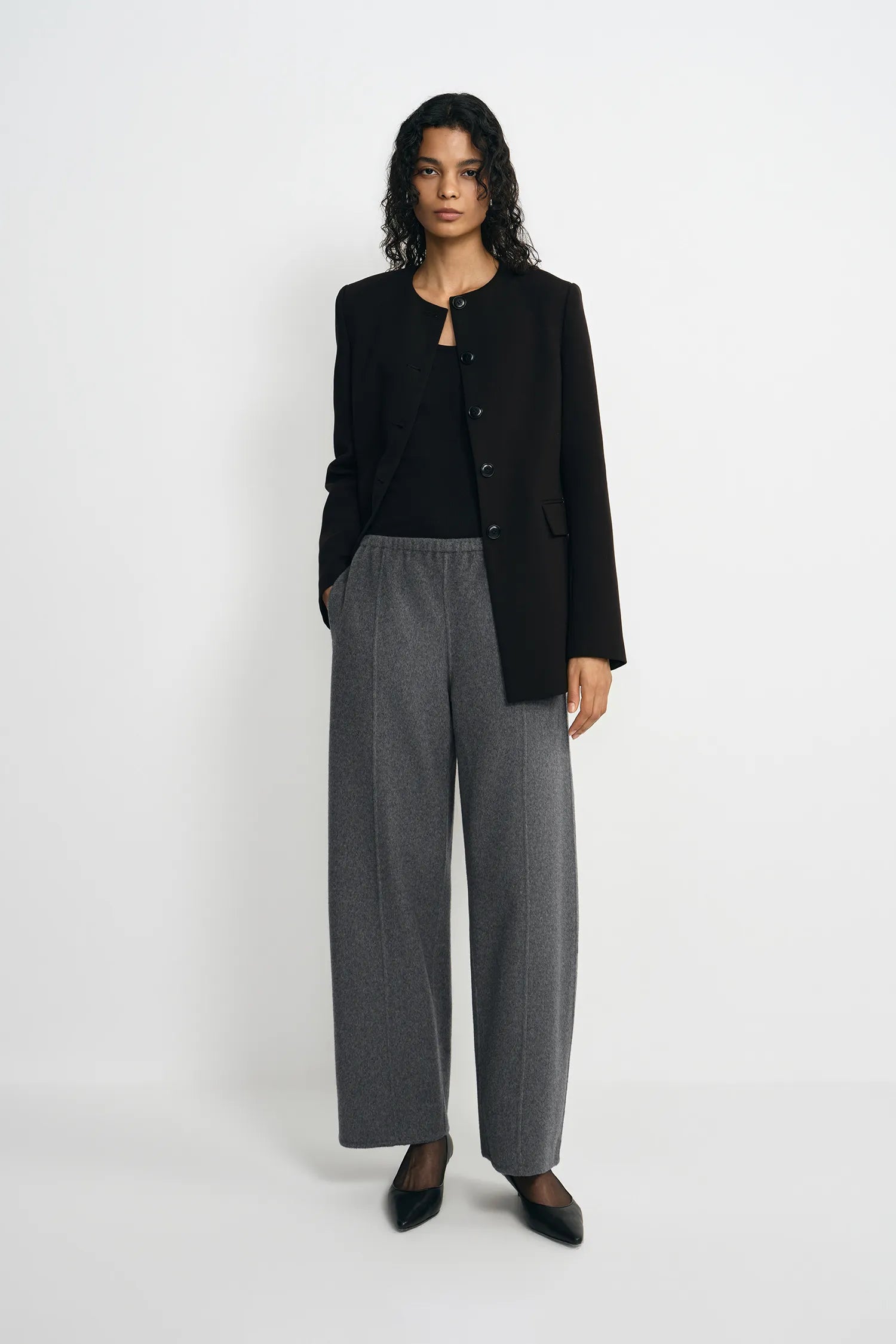 A model stands against a light gray studio background wearing a black cropped button-front jacket over a black top, paired with wide-leg gray trousers and black pointed shoes.