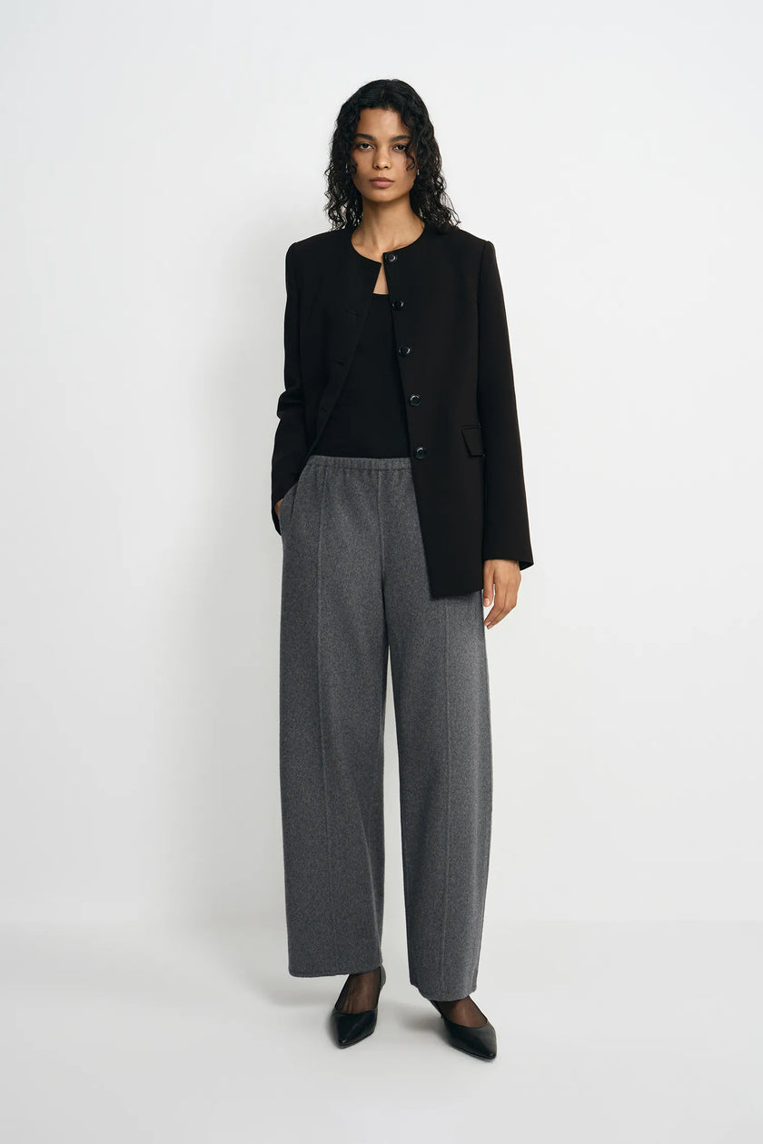 A model stands against a light gray studio background wearing a black cropped button-front jacket over a black top, paired with wide-leg gray trousers and black pointed shoes.