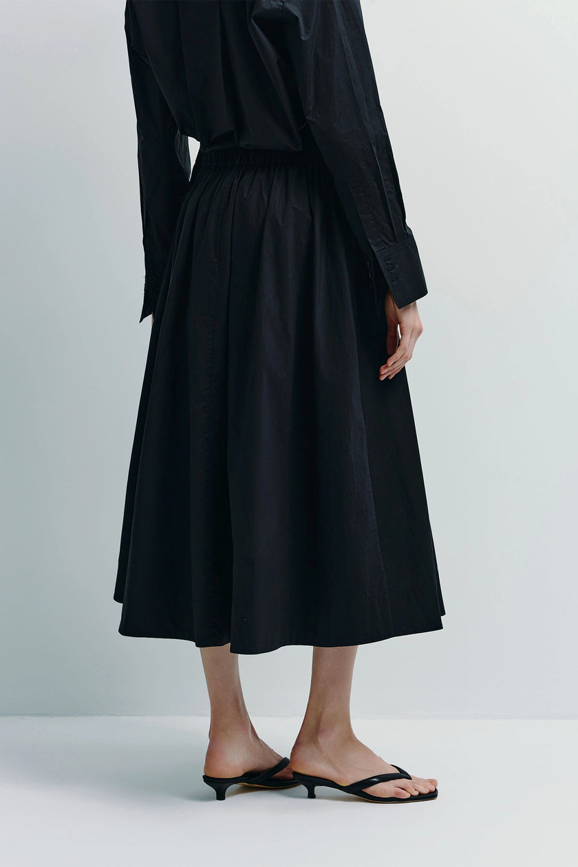 Black Andra Poplin Midi Skirt, flowy silhouette, elastic waist, paired with a black shirt and sandals