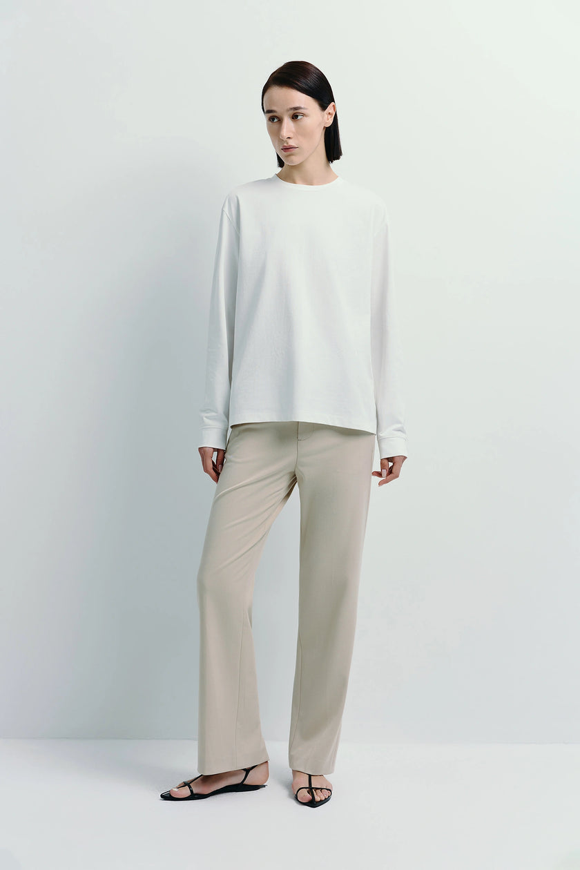 Anza Long Sleeve Top in white, paired with beige trousers, offering a relaxed, layered look.