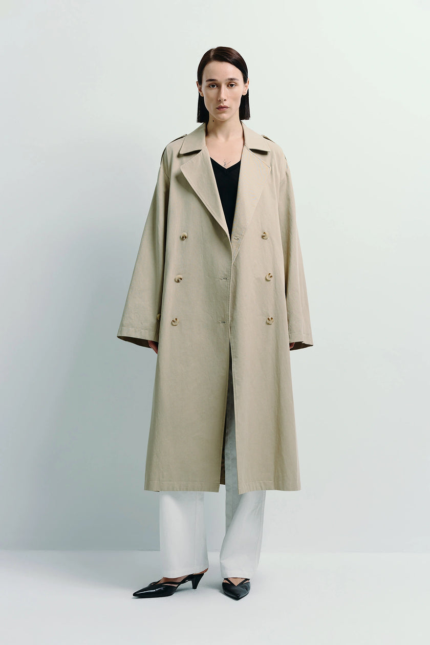 Beige Balham Trench Coat, double-breasted design. Model wearing black top and white pants.