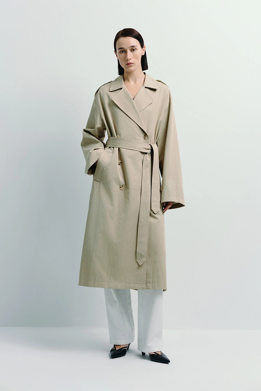 Beige Balham Trench Coat, double-breasted with belt, worn with white pants by Rue Sophie.