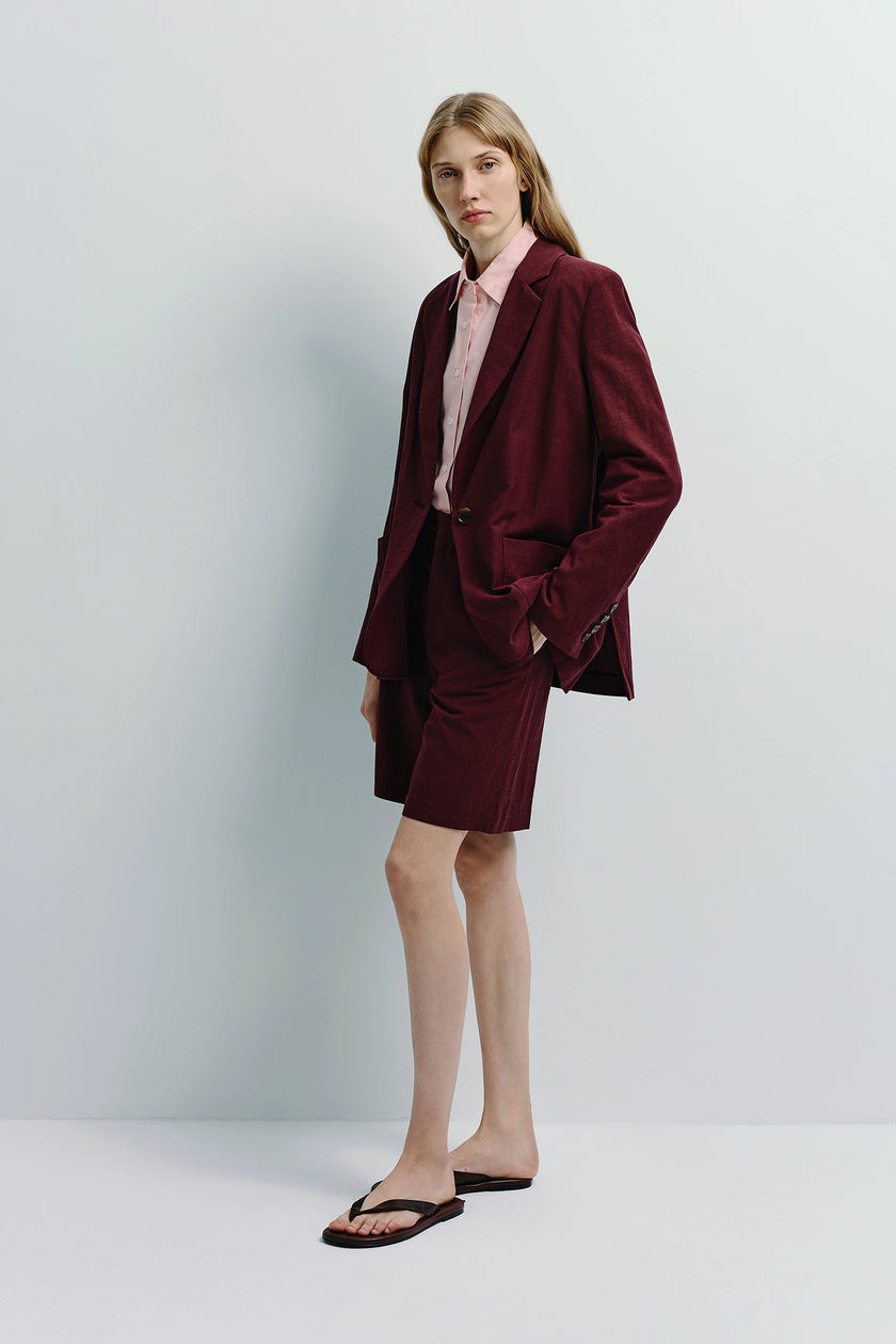 Woman in maroon linen Bermuda shorts suit, pink shirt, and sandals. Versatile outfit.