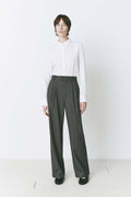 Model wearing pleated front straight leg trousers with an extended tab closure, styled with a white button-up shirt, showcasing a tailored fit and refined silhouette for a sophisticated, professional look.