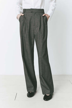 Model wearing pleated front straight leg trousers with an extended tab closure, styled with a white button-up shirt, showcasing a tailored fit and refined silhouette for a sophisticated, professional look.