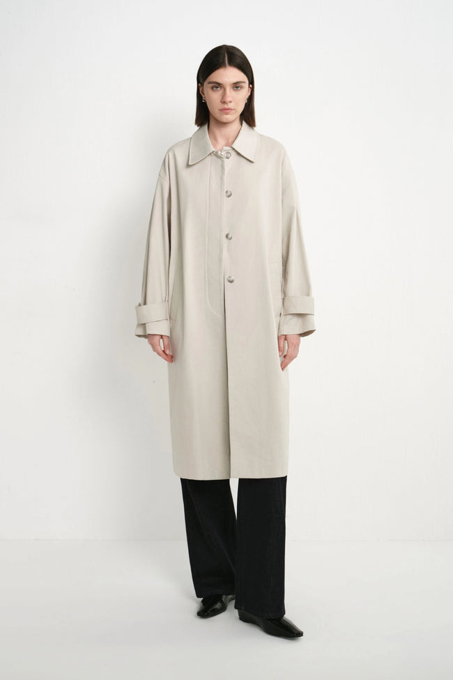 A model stands against a plain white studio background wearing a long, light beige trench coat with a collar and front buttons, paired with wide-leg black trousers and black shoes.