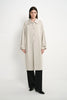 A model stands against a plain white studio background wearing a long, light beige trench coat with a collar and front buttons, paired with wide-leg black trousers and black shoes.