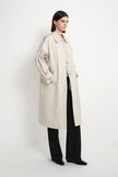 A model stands in a studio wearing a long, oversized light beige trench coat over black wide-leg trousers and black shoes, photographed against a plain white background.