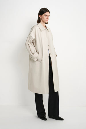 A model stands in a studio wearing a long, oversized light beige trench coat over black wide-leg trousers and black shoes, photographed against a plain white background.