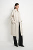 A model stands in a studio wearing a long, oversized light beige trench coat over black wide-leg trousers and black shoes, photographed against a plain white background.