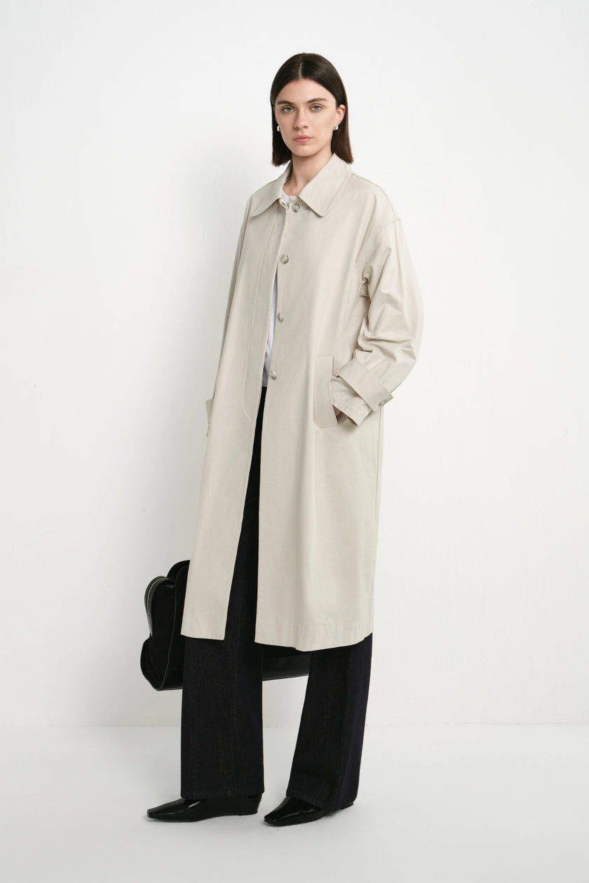 A model stands against a plain white studio background wearing a long light beige trench coat over a white collared shirt and black wide-leg trousers, with black shoes and a black bag.