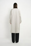 A model viewed from the back wearing a light beige long trench coat over black wide-leg trousers, set against a plain white studio background.