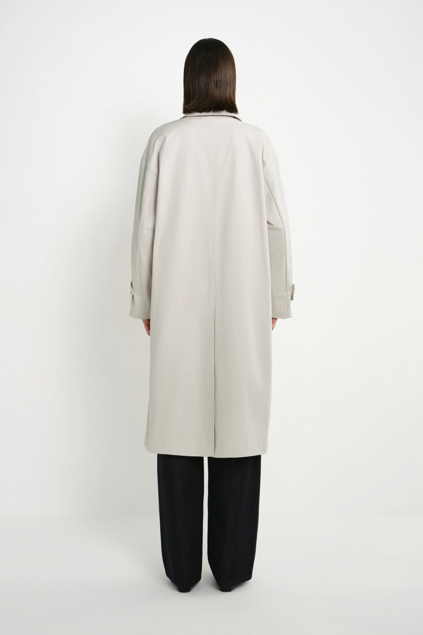 A model viewed from the back wearing a light beige long trench coat over black wide-leg trousers, set against a plain white studio background.