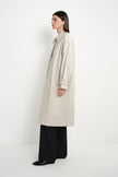 A model is shown in profile wearing a long, light beige trench coat over black wide-leg trousers and black shoes, standing against a plain white studio background.