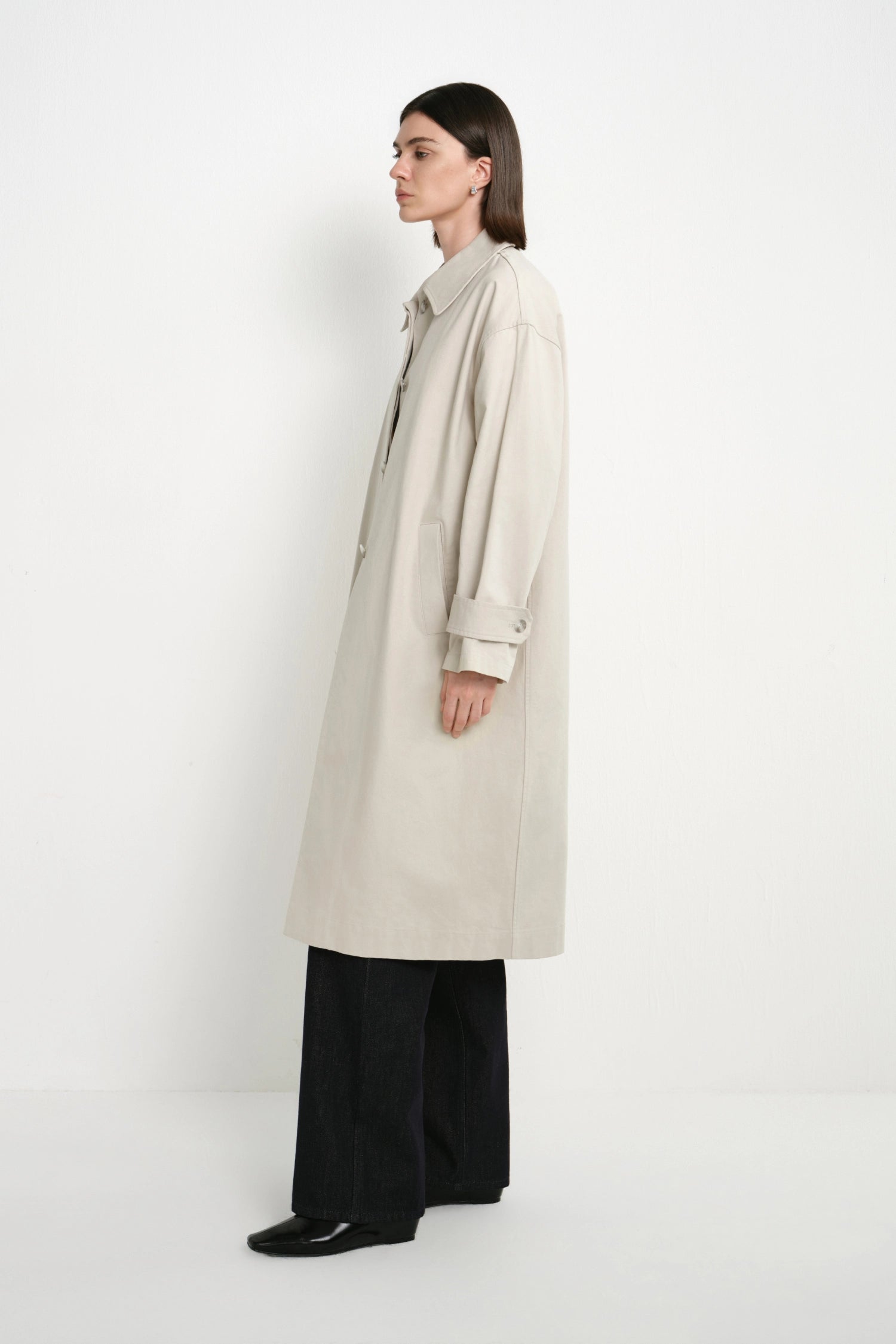 A model is shown in profile wearing a long, light beige trench coat over black wide-leg trousers and black shoes, standing against a plain white studio background.