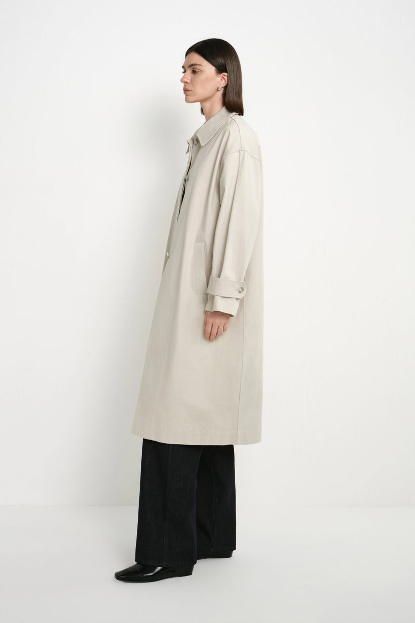 A model is shown in profile wearing a long, light beige trench coat over black wide-leg trousers and black shoes, standing against a plain white studio background.