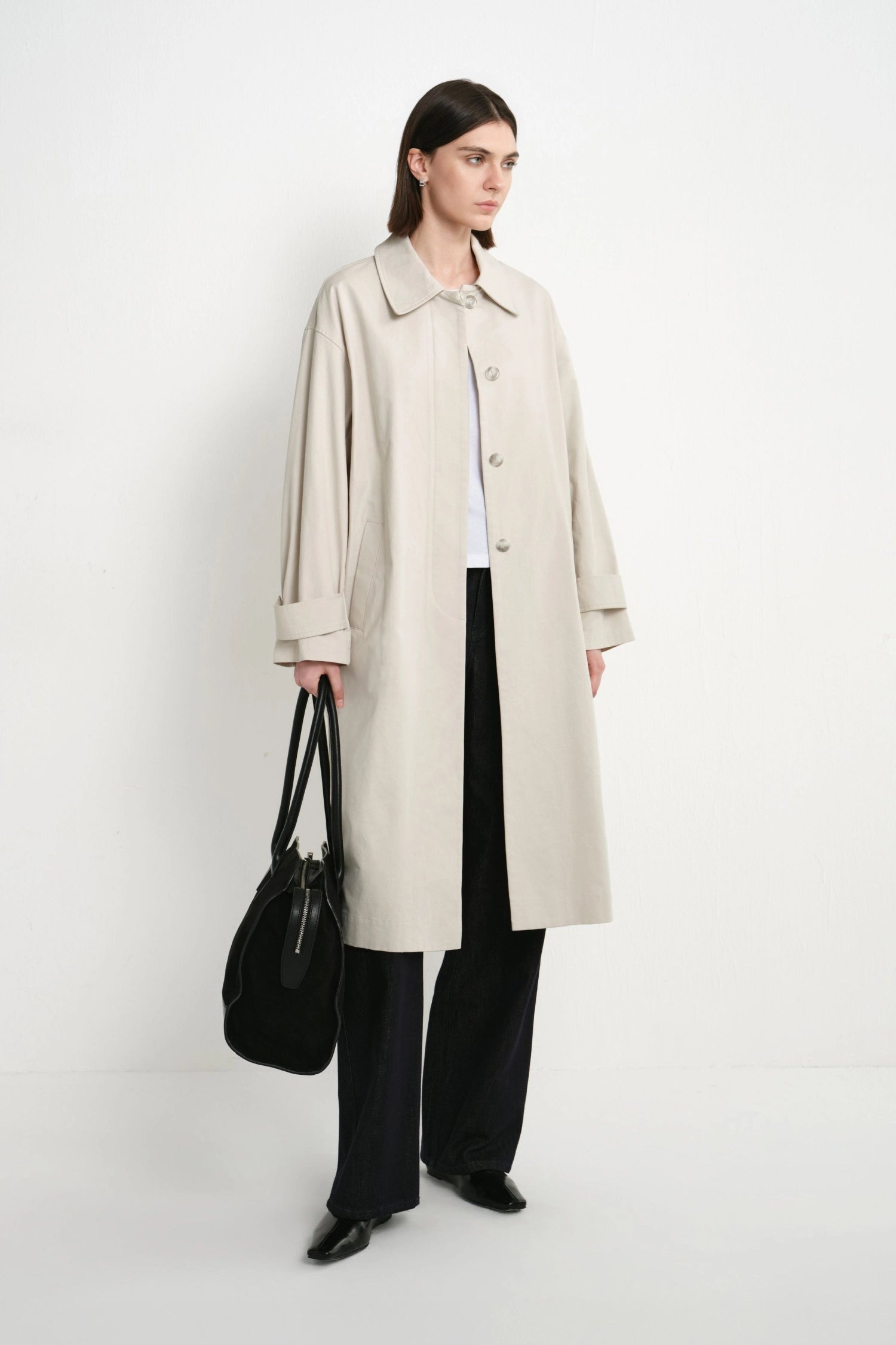 A model stands against a plain white studio background wearing a long light beige trench coat over a white top and black wide-leg trousers, while holding a black handbag.