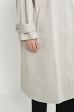 A close-up of a person wearing a light beige trench coat, showing the sleeve with a button cuff and a side pocket, paired with black pants against a plain white background.