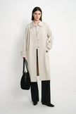 A model stands against a plain white studio background wearing a long light beige trench coat over a white top and black wide-leg trousers, while holding a black shoulder bag.