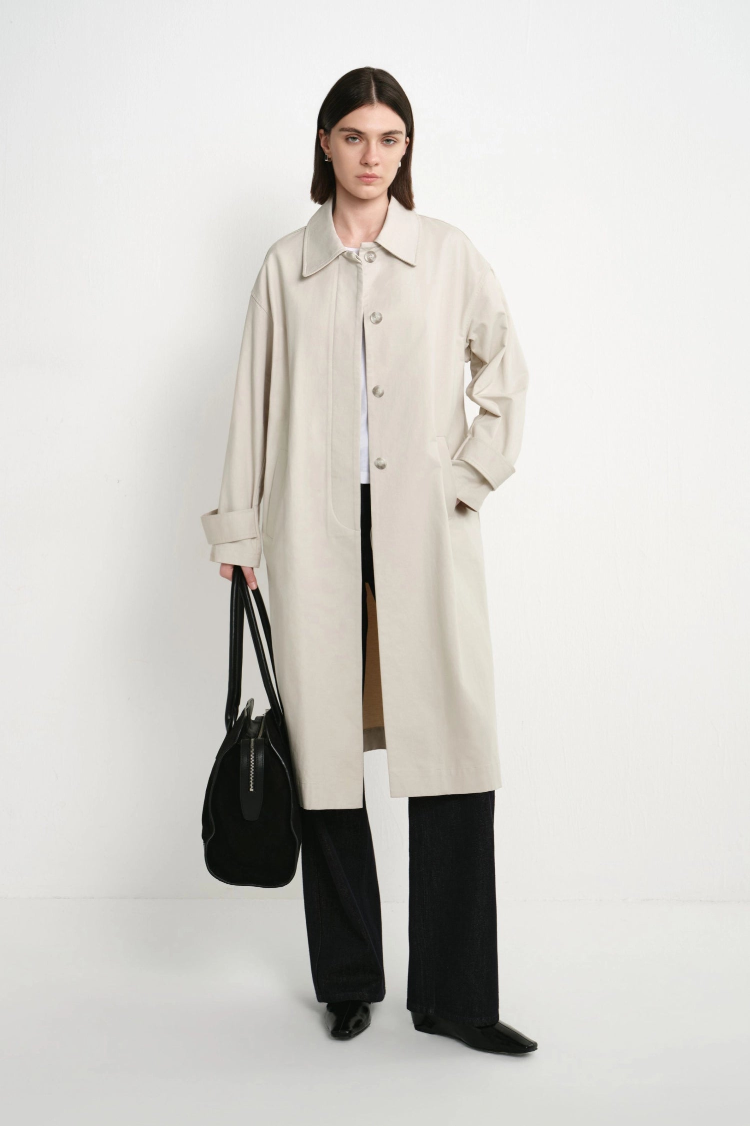 A model stands against a plain white studio background wearing a long light beige trench coat over a white top and black wide-leg trousers, while holding a black shoulder bag.