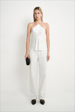 A woman modeling a white sleeveless halter top and matching wide-leg trousers, standing against a plain white studio background and holding a small black clutch.