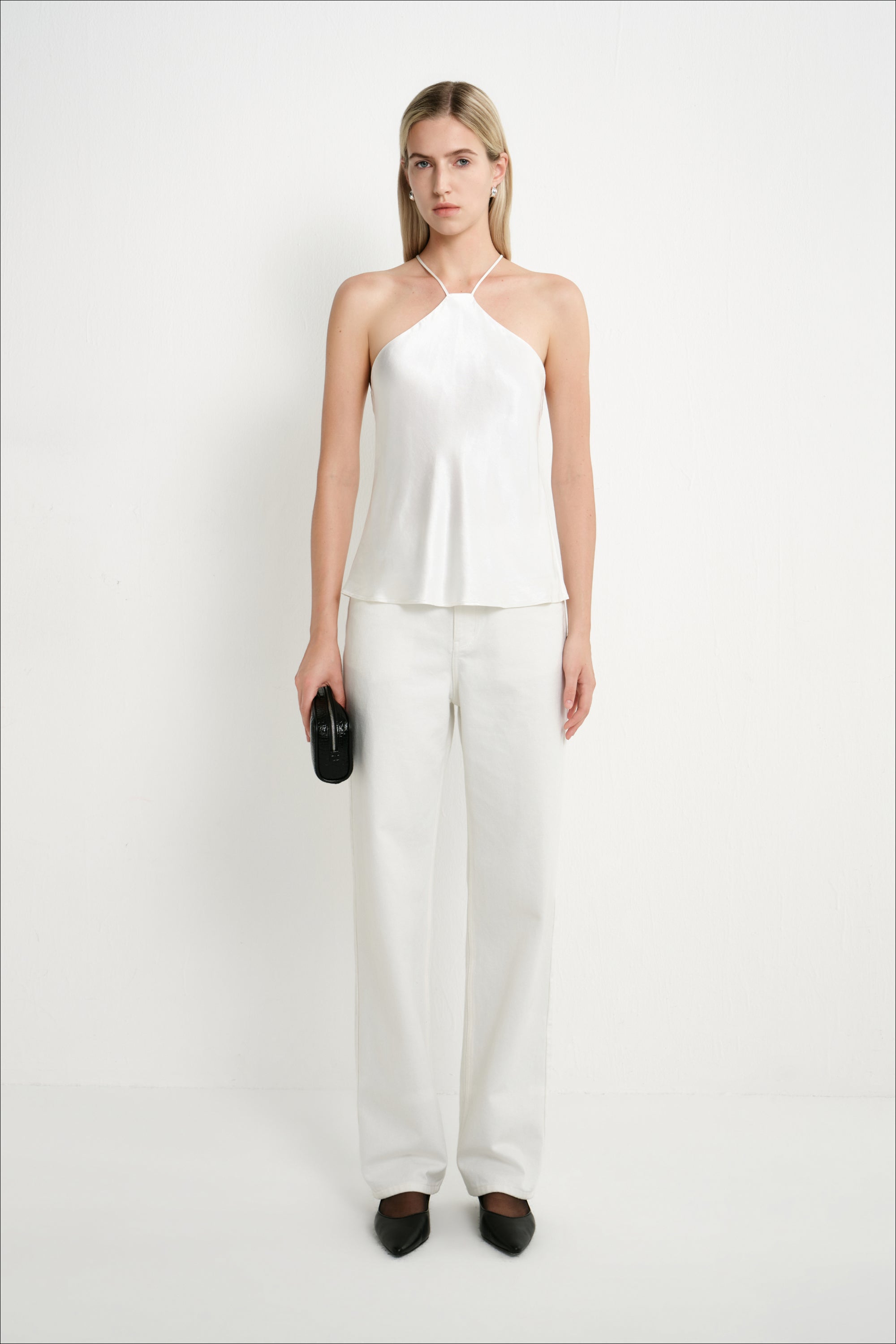 A woman modeling a white sleeveless halter top and matching wide-leg trousers, standing against a plain white studio background and holding a small black clutch.