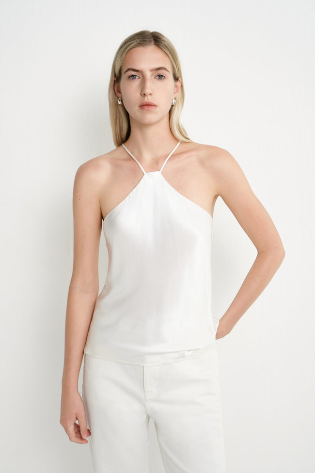 A blonde model wearing a white halter-neck camisole top and matching white trousers, standing against a plain white studio background.