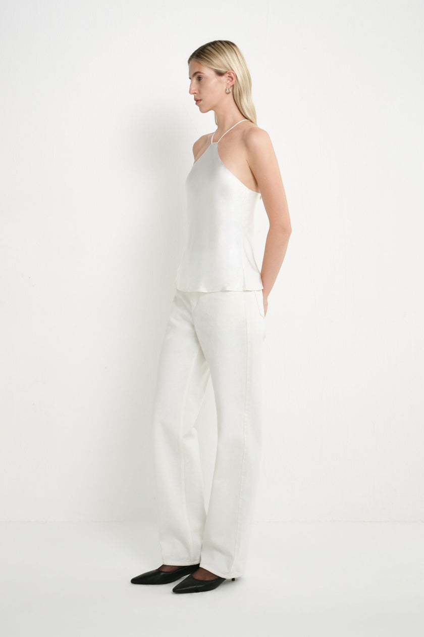 A woman stands in profile wearing a white sleeveless camisole top and matching white wide-leg trousers, paired with black pointed flats, against a plain white studio background.