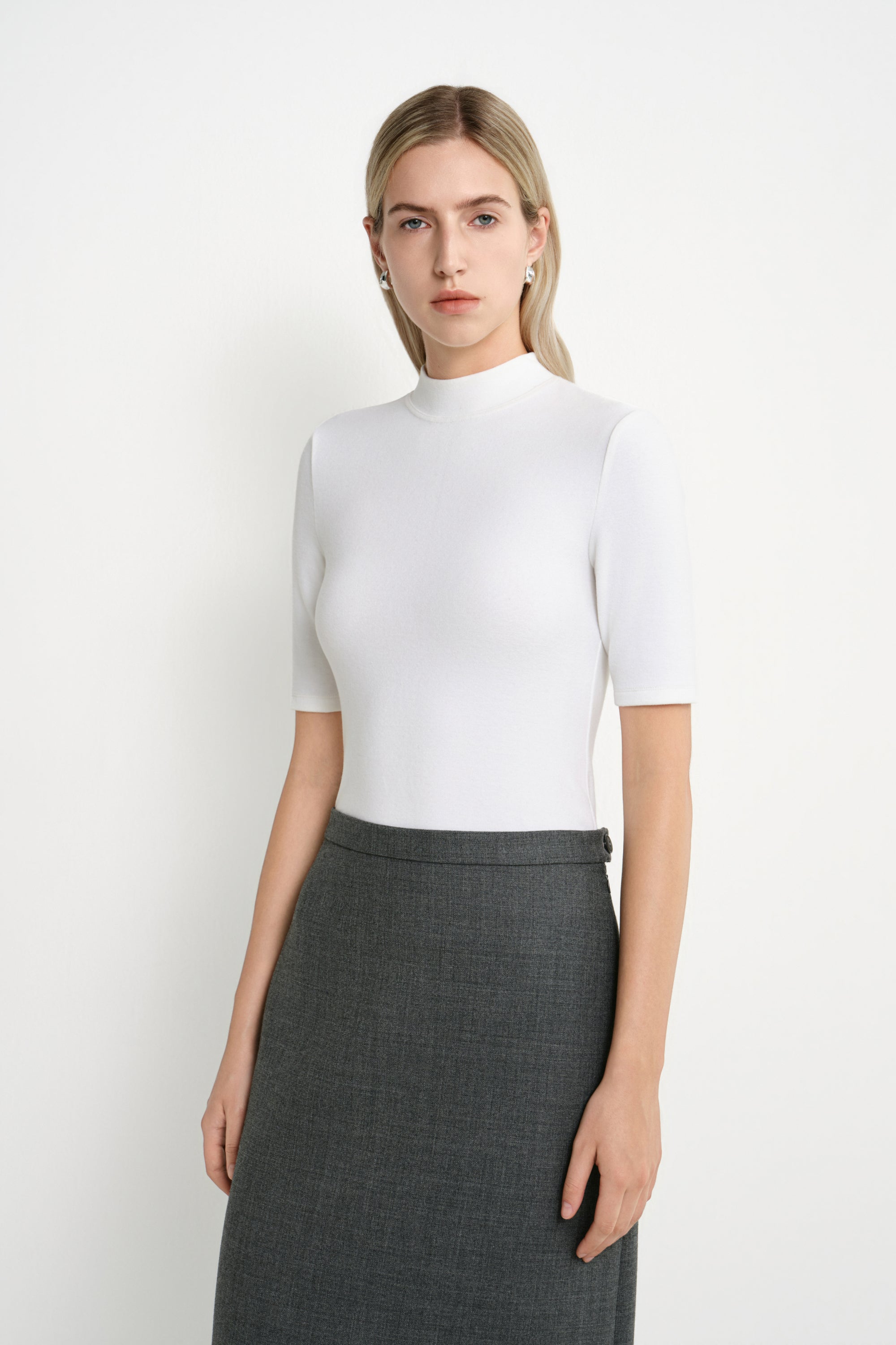 A model wearing the Rue Sophie Chabot Mock Neck Top in White with a streamlined high-waisted silhouette and wide structured hem, styled with black knee-high boots, front view.