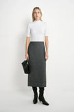 A woman stands against a plain white studio background wearing a fitted white short-sleeve mock-neck top, a dark gray knee-length skirt, black knee-high socks, and black heeled boots, while holding a black clutch.