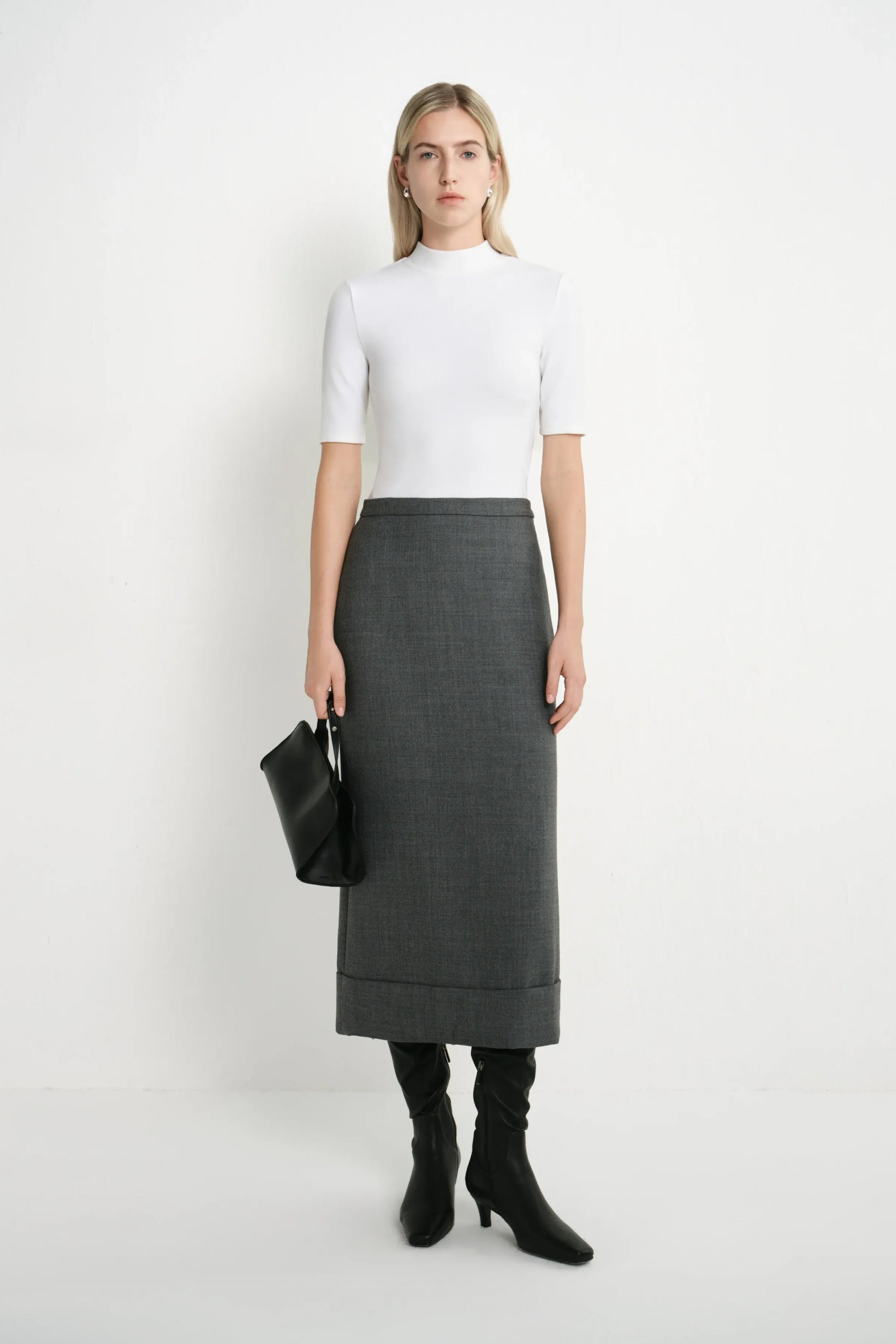 A woman stands against a plain white studio background wearing a fitted white short-sleeve mock-neck top, a dark gray knee-length skirt, black knee-high socks, and black heeled boots, while holding a black clutch.