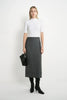 A woman stands against a plain white studio background wearing a fitted white short-sleeve mock-neck top, a dark gray knee-length skirt, black knee-high socks, and black heeled boots, while holding a black clutch.