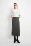 A woman stands against a plain white studio background wearing a white short-sleeve mock-neck top, a dark gray knee-length skirt, and black heeled boots.