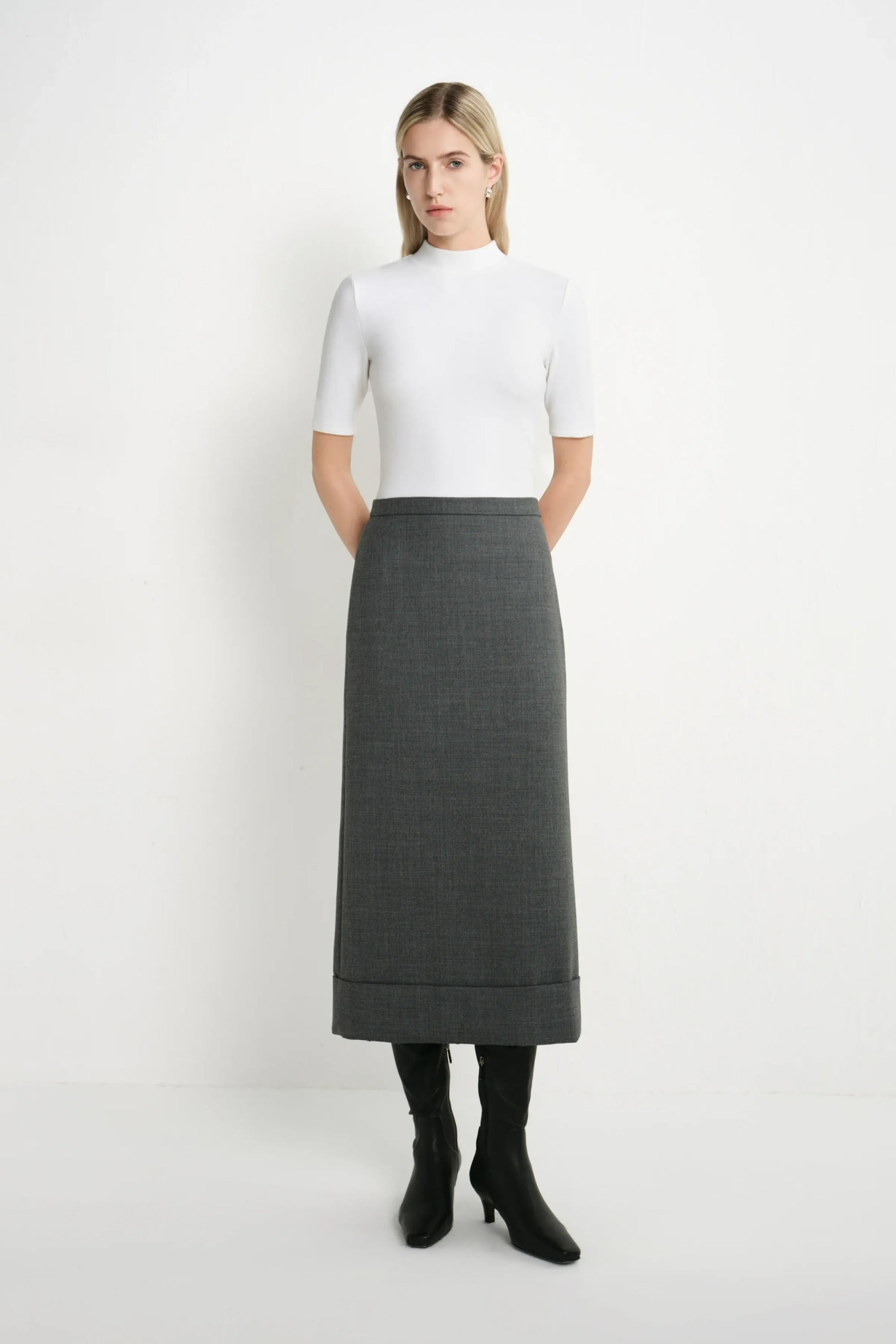 A woman stands against a plain white studio background wearing a white short-sleeve mock-neck top, a dark gray knee-length skirt, and black heeled boots.