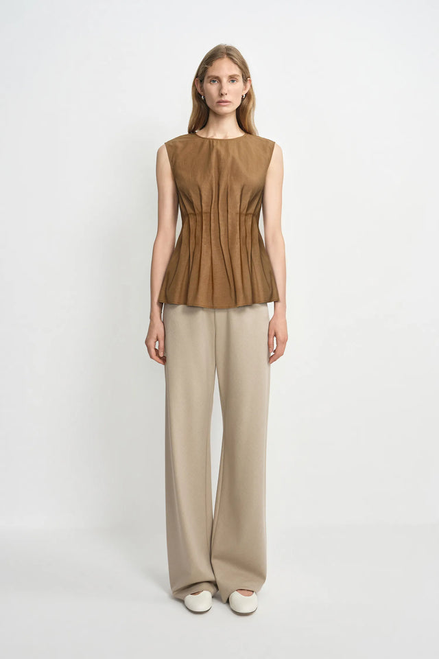 A woman stands against a plain light gray studio background wearing a sleeveless brown peplum-style top with gathered detailing and beige wide-leg pants.