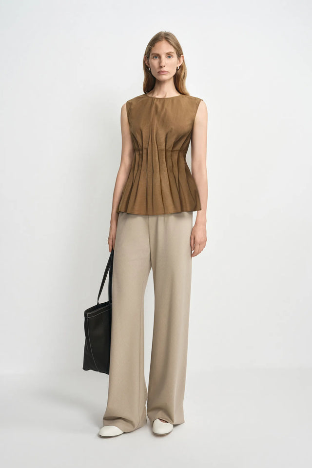A model stands against a light gray studio background wearing a sleeveless brown peplum top, beige wide-leg trousers, white pointed shoes, and holding a black tote bag.