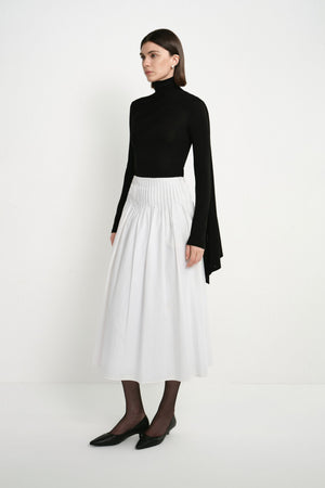 A model wearing the Rue Sophie Piquenique Pintuck Skirt in white, highlighting the high pintucked waistband, gathered volume, side pockets, and flowing midi length, three-quarter view.