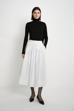A model wearing the Rue Sophie Piquenique Pintuck Skirt in white, featuring a wide pintucked waistband, gathered volume, and a flowing midi-length silhouette, front view.