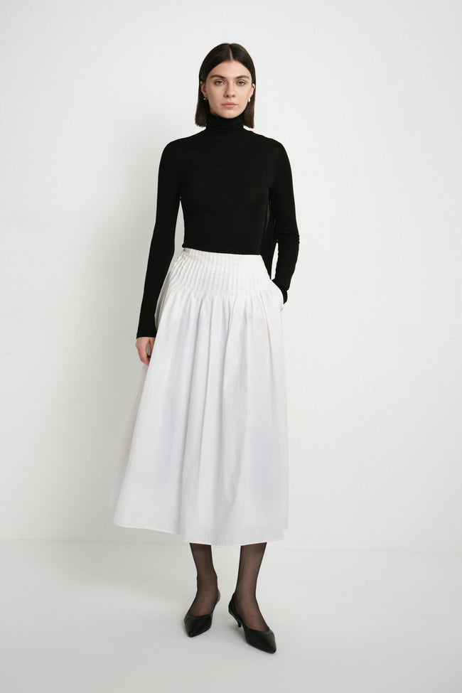 A model wearing the Rue Sophie Piquenique Pintuck Skirt in white, featuring a wide pintucked waistband, gathered volume, and a flowing midi-length silhouette, front view.