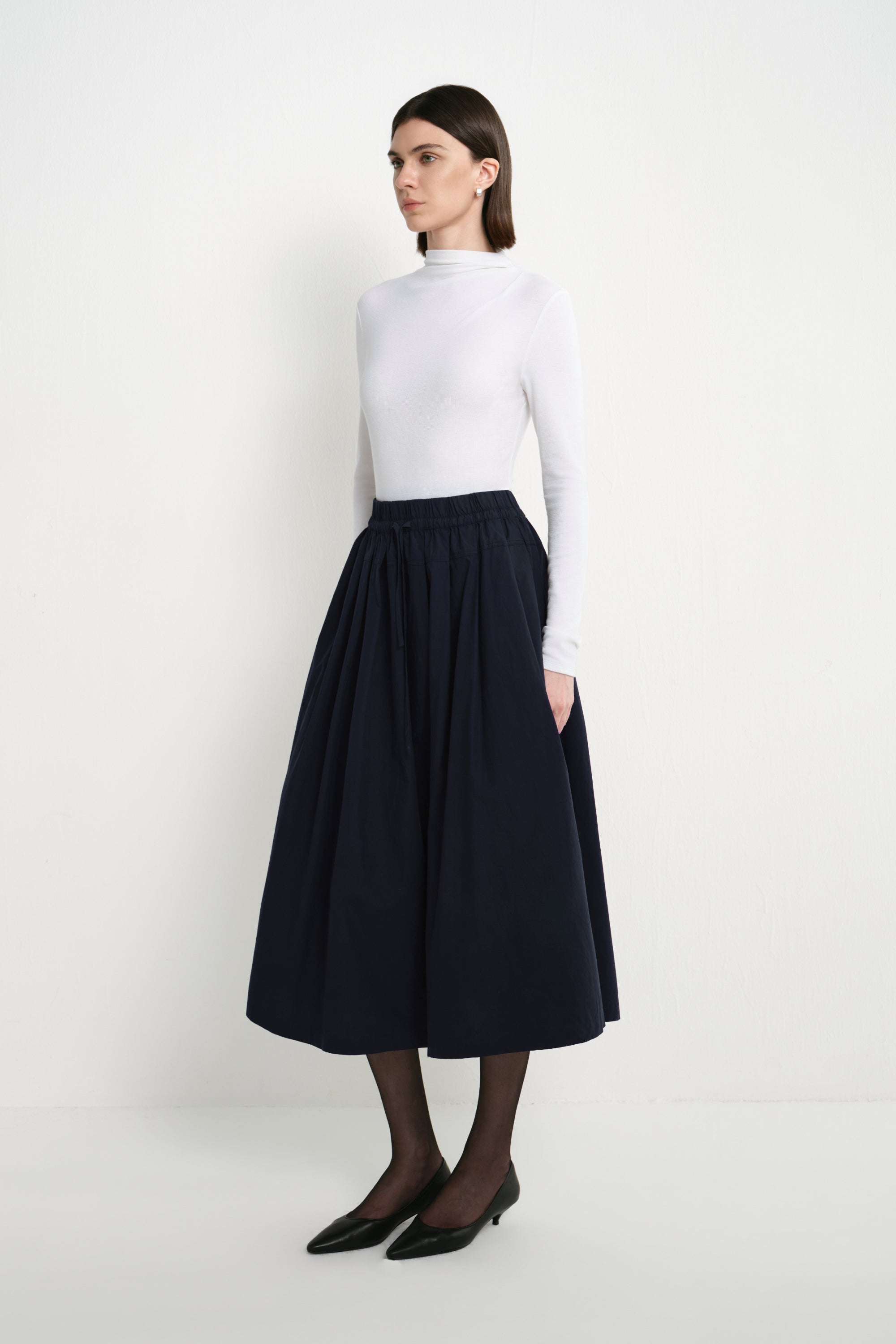A model wearing the Rue Sophie Panier Drawstring Waist Pleated Skirt in Navy, showing its gathered volume and midi-length silhouette from a side angle.